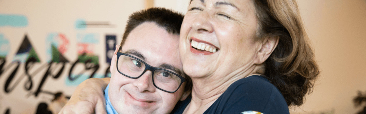 Woman hugging a young man with Down syndrome wearing glasses. Both are smiling. 